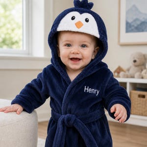 May include: A navy blue plush bathrobe featuring a hood designed to look like a penguin with a white face, black eyes, and an orange beak. The robe has long sleeves, a tie belt at the waist, and the name Henry embroidered in white on the chest.