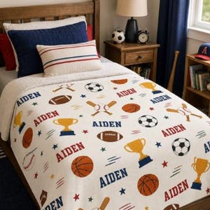 May include: A white blanket featuring a repeating pattern of sports equipment including footballs, basketballs, soccer balls, baseballs with bats, and gold trophies, interspersed with the name Aiden in blue and red text on a wooden bed frame.
