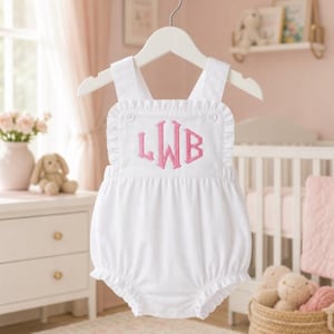 Könnte beinhalten: Ein ärmelloser weißer Strampler mit gerüschten Schulterträgern und einem gerüschten Ausschnitt. Die Vorderseite ist mit einem rosa Monogramm mit den Buchstaben LWB bestickt. Das Kleidungsstück hat eine geraffte Taille und Beinöffnungen mit Druckknöpfen.