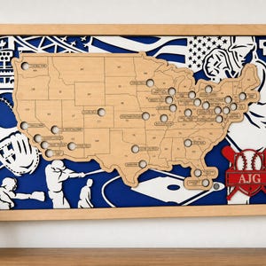 May include: A rectangular wooden wall art piece featuring a layered map of the United States with cutouts for baseball stadiums. The design includes white and blue silhouettes of baseball players, a glove, and a flag. A red emblem displays the letters AJG.