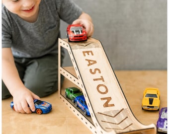 May include: A wooden toy car ramp featuring the name EASTON engraved on the surface. The ramp has two levels for storing small die-cast toy cars. A red car is positioned at the top, while a green and a blue car are parked on the lower level.