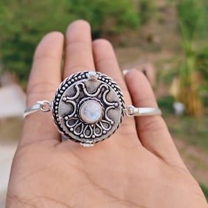 Puede incluir: Una pulsera de plata con una pieza central circular de acabado oxidado y una piedra lunar iridiscente en el centro. El punto focal redondo muestra intrincados patrones en relieve y un borde de cuerda trenzada, unido a una banda lisa.