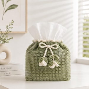 May include: A sage green crochet tissue box cover featuring a textured stitch pattern, a cream-coloured scalloped top edge, and a decorative cream-coloured drawstring tie with two small, dangling crochet flower embellishments at the centre front.