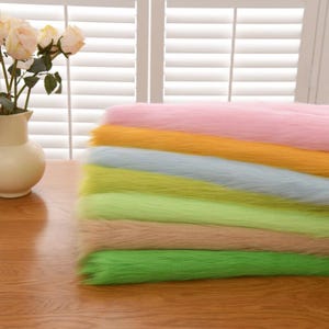 Könnte beinhalten: Ein Stapel gefalteter Kunstfellstoffstücke in verschiedenen Farben, darunter Rosa, Gelb, Hellblau, Limettengrün, Hellbraun und leuchtendes Grün, neben einer weißen Keramikvase mit mehreren blassrosa künstlichen Rosenblüten.