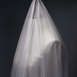 Może przedstawiać: Półprzezroczysty, biały welon z tkaniny narzucony na formę. Lekki materiał tworzy miękkie, pionowe fałdy i marszczenia, zasłaniając kształt pod spodem, jednocześnie przepuszczając światło przez delikatną, warstwową powierzchnię tekstylną.
