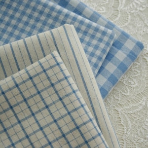 Puede incluir: Tres piezas de tela dobladas con patrones en azul y blanco. Una pieza tiene un estampado de cuadros vichy en azul y blanco, otra tiene finas rayas verticales azules sobre fondo crema y la tercera tiene un patrón de cuadrícula azul sobre tela crema.