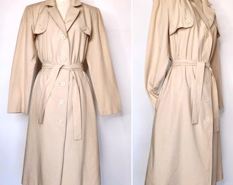 May include: A tan trench coat with a belted waist and button-down closure. The coat features a classic collar, shoulder epaulets, and long sleeves. The coat is displayed on a mannequin, showcasing its tailored design and overall silhouette.