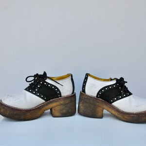 May include: A pair of white and black saddle shoes with a thick platform sole. The shoes have a black suede toe and a white leather upper. The shoes are worn and have dirt on the soles.