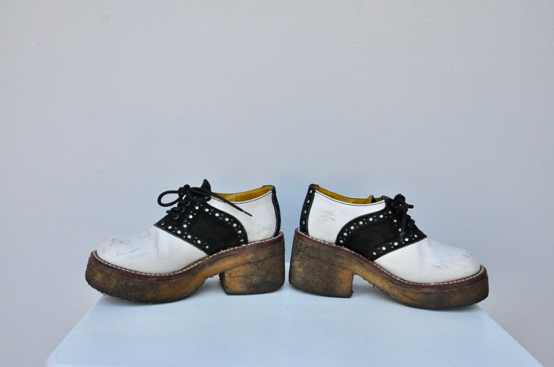 May include: A pair of white and black saddle shoes with a thick platform sole. The shoes have a black suede toe and a white leather upper. The shoes are worn and have dirt on the soles.