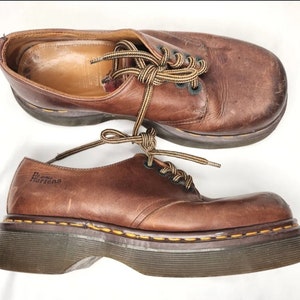 Può includere: Un paio di scarpe marroni in pelle Dr. Martens con lacci e suola spessa nera. Le scarpe hanno un aspetto usurato e sono legate con lacci marroni.