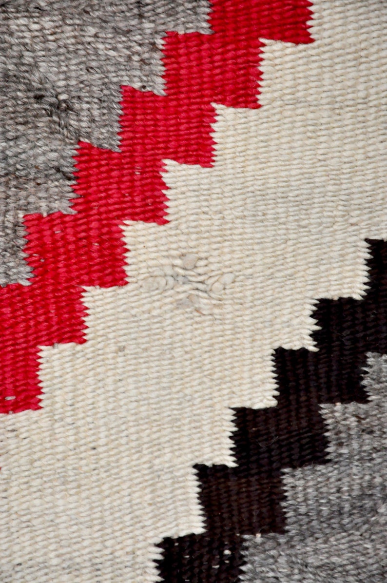 May include: Close-up of a woven rug with a geometric pattern in shades of red, white, black, and gray.