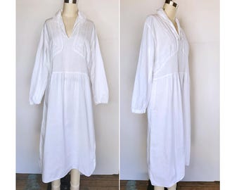 May include: A white cotton dress with long sleeves and a v-neck collar. The dress has a gathered skirt and a relaxed fit.