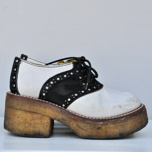 May include: White and black leather platform saddle shoes with a lace-up closure. The shoes have a thick, worn platform sole.