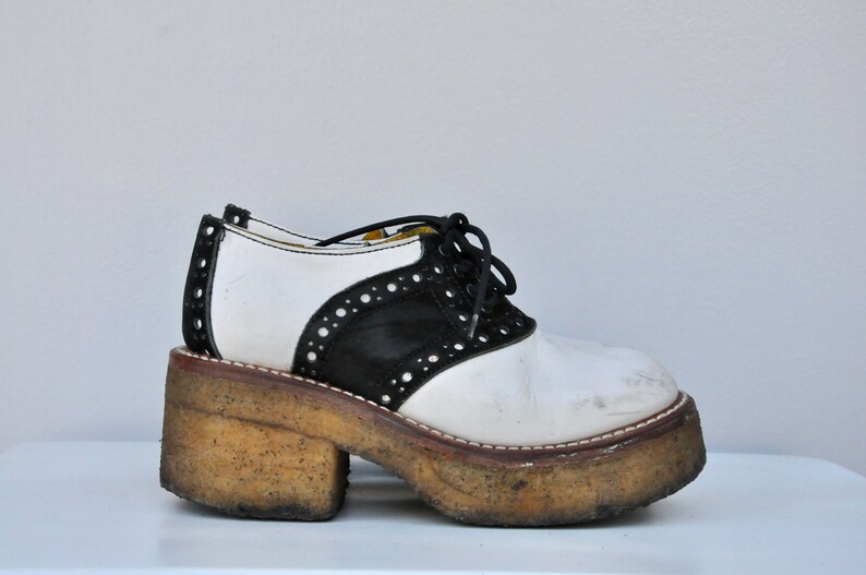May include: White and black leather platform saddle shoes with a lace-up closure. The shoes have a thick, worn platform sole.