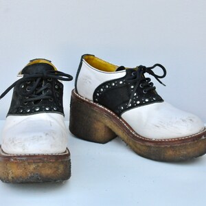 May include: A pair of white and black platform saddle shoes with black laces. The shoes have a thick platform sole and are worn.