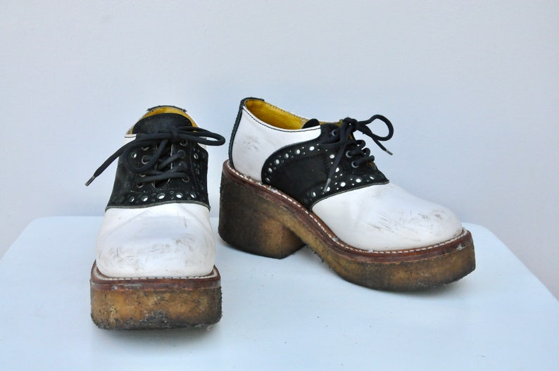 May include: A pair of white and black platform saddle shoes with black laces. The shoes have a thick platform sole and are worn.