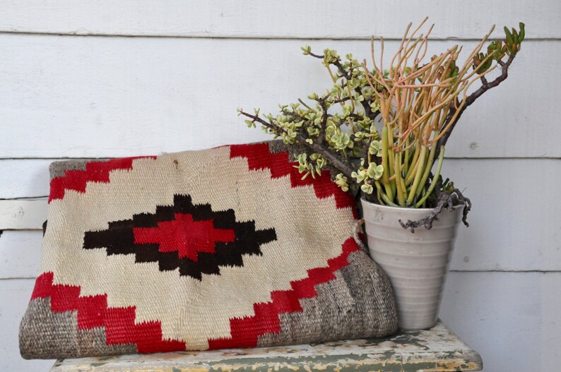 May include: A white, red, and brown woven rug with a geometric pattern. The rug is sitting on a wooden surface with a potted plant in the background.