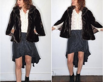 May include: A brown faux fur cape with a high collar and long sleeves. The cape is worn over a white blouse with a lace pattern. The person is wearing a black high-low skirt.