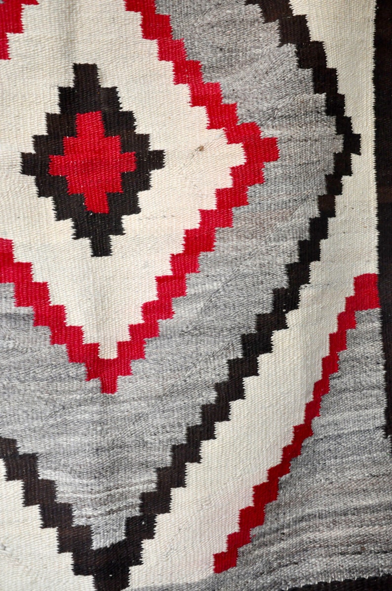 May include: A close-up of a woven rug with a geometric pattern in shades of red, black, white, and gray.