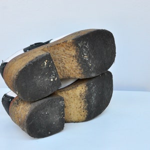 May include: A pair of black and white platform shoes with a thick wooden sole. The shoes have a buckle closure and are worn.