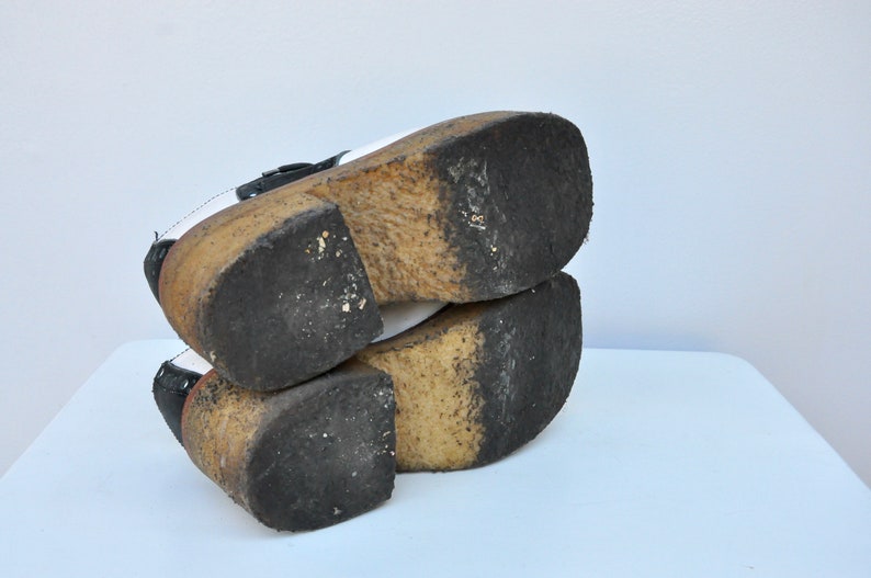 May include: A pair of black and white platform shoes with a thick wooden sole. The shoes have a buckle closure and are worn.