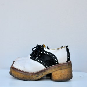 May include: White and black platform saddle shoes with a thick, chunky sole. The shoes have a black suede panel and white leather panels with black stitching and white laces.
