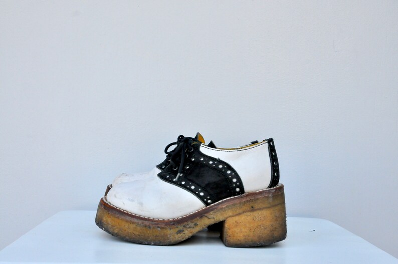 May include: White and black platform saddle shoes with a thick, chunky sole. The shoes have a black suede panel and white leather panels with black stitching and white laces.