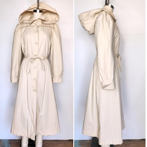 May include: A long, cream-colored raincoat with a hood and a tie belt. The raincoat has a button-up front and long sleeves.