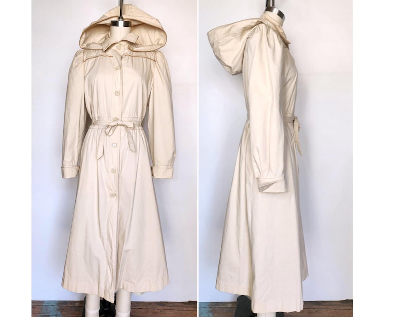 May include: A long, cream-colored raincoat with a hood and a tie belt. The raincoat has a button-up front and long sleeves.