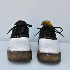 May include: A pair of white and black saddle shoes with black laces. The shoes have a platform sole and are worn.