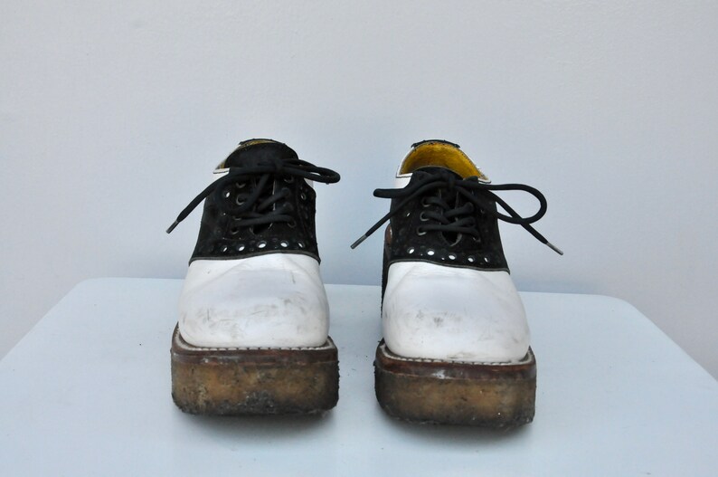 May include: A pair of white and black saddle shoes with black laces. The shoes have a platform sole and are worn.