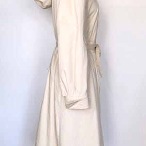 May include: A long, cream-colored hooded jacket with a cinched waist and a tie belt. The jacket has a relaxed fit and a flowing skirt.