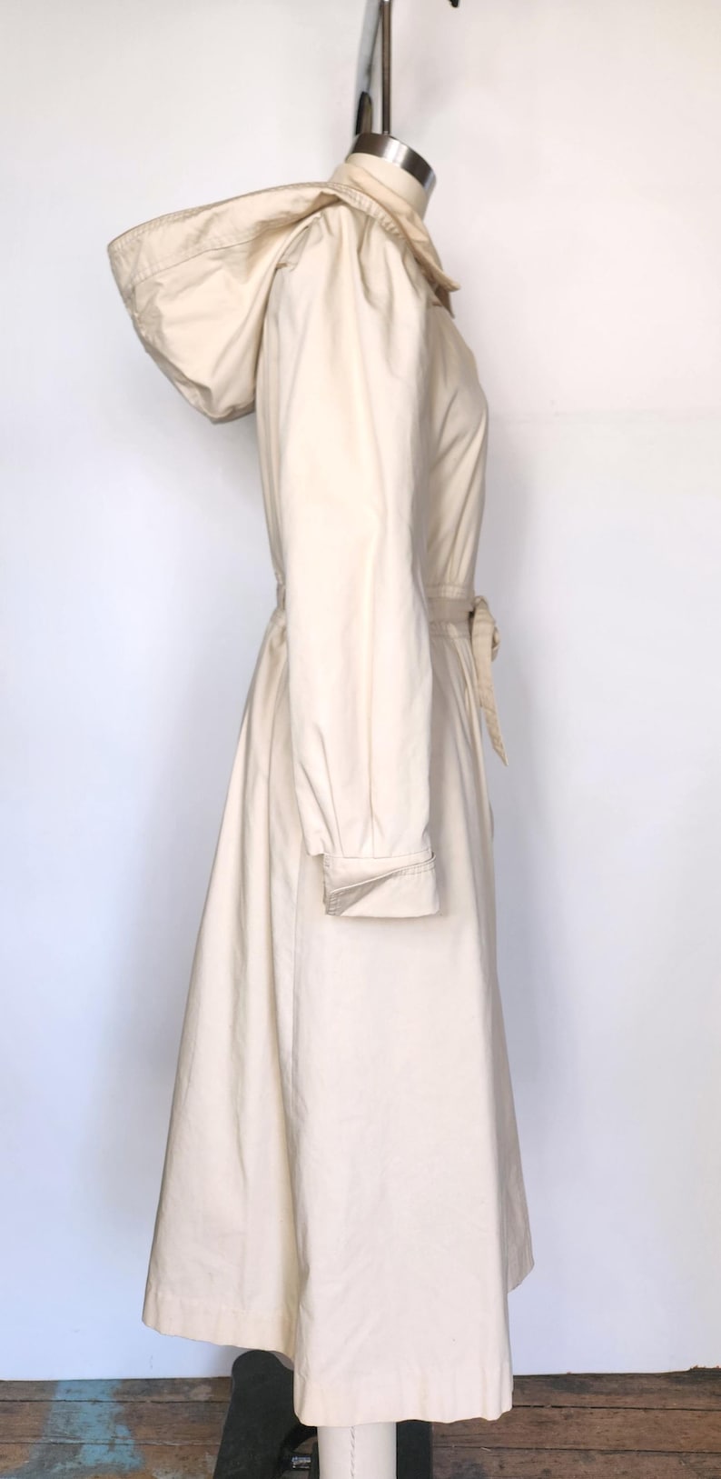 May include: A long, cream-colored hooded jacket with a cinched waist and a tie belt. The jacket has a relaxed fit and a flowing skirt.