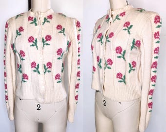 May include: Cream-coloured, button-up cardigan sweater with a floral pattern. The sweater features pink roses with green stems and leaves. The sleeves and body have a ribbed texture.
