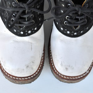 May include: A pair of white and black leather saddle shoes with black laces. The shoes have a white leather upper and a black leather trim around the toe and heel. The laces are tied in a bow at the top of the shoes.