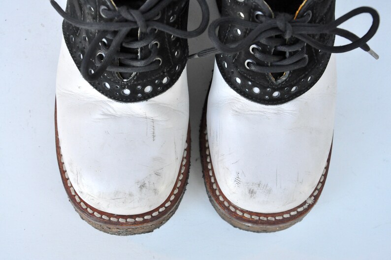 May include: A pair of white and black leather saddle shoes with black laces. The shoes have a white leather upper and a black leather trim around the toe and heel. The laces are tied in a bow at the top of the shoes.
