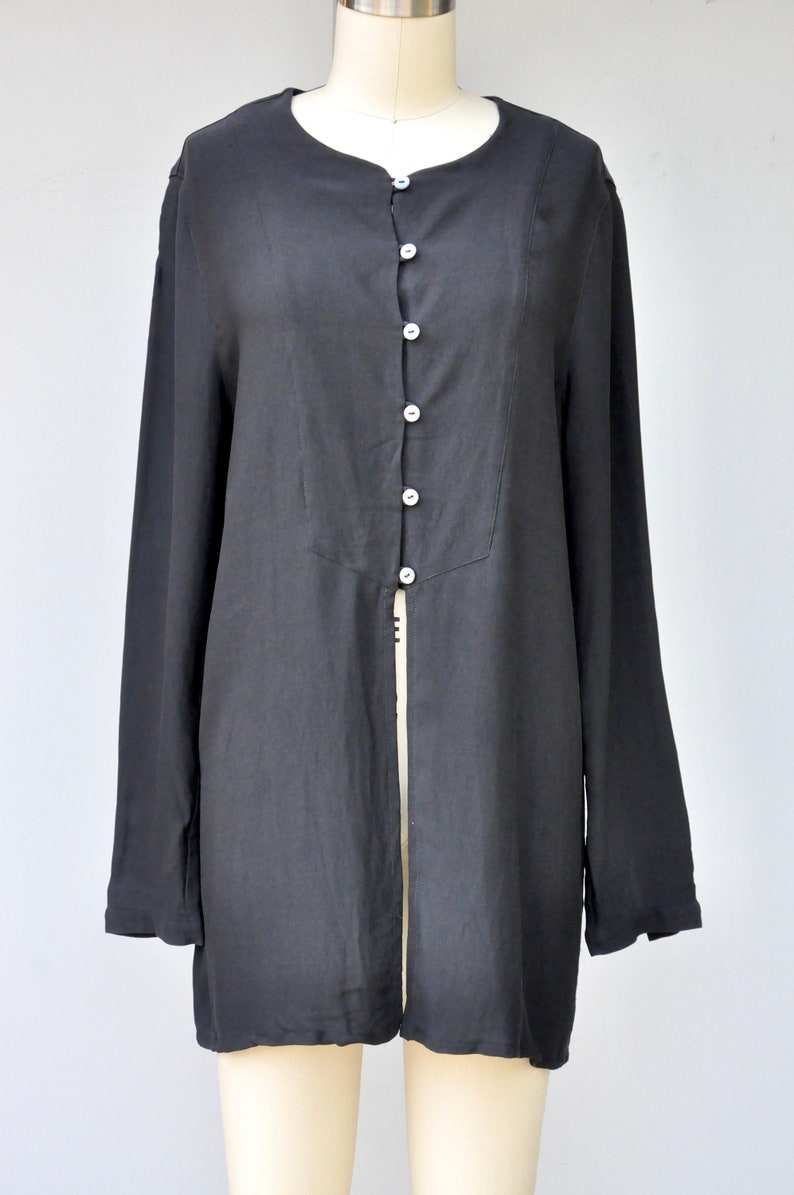 May include: A long-sleeved black button-up shirt with a center front slit. The shirt has a round neckline and a relaxed fit.