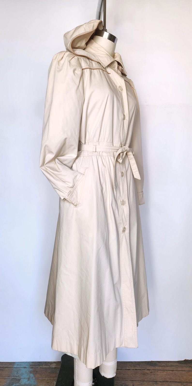 May include: A long-sleeved, cream-colored hooded jacket with a button-up front and a tie belt. The jacket has a relaxed fit and a slightly flared skirt.