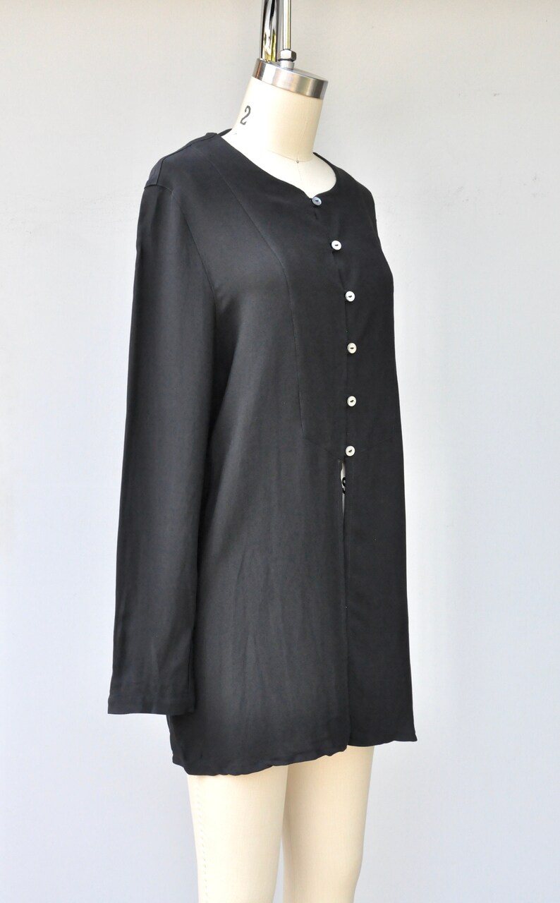 May include: A black long-sleeved button-up shirt with a round neckline. The shirt is made of a smooth, silky fabric and has a slightly loose fit.
