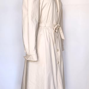 May include: A long, off-white hooded trench coat with a belt and button closure. The coat has a slightly flared skirt and a relaxed fit.