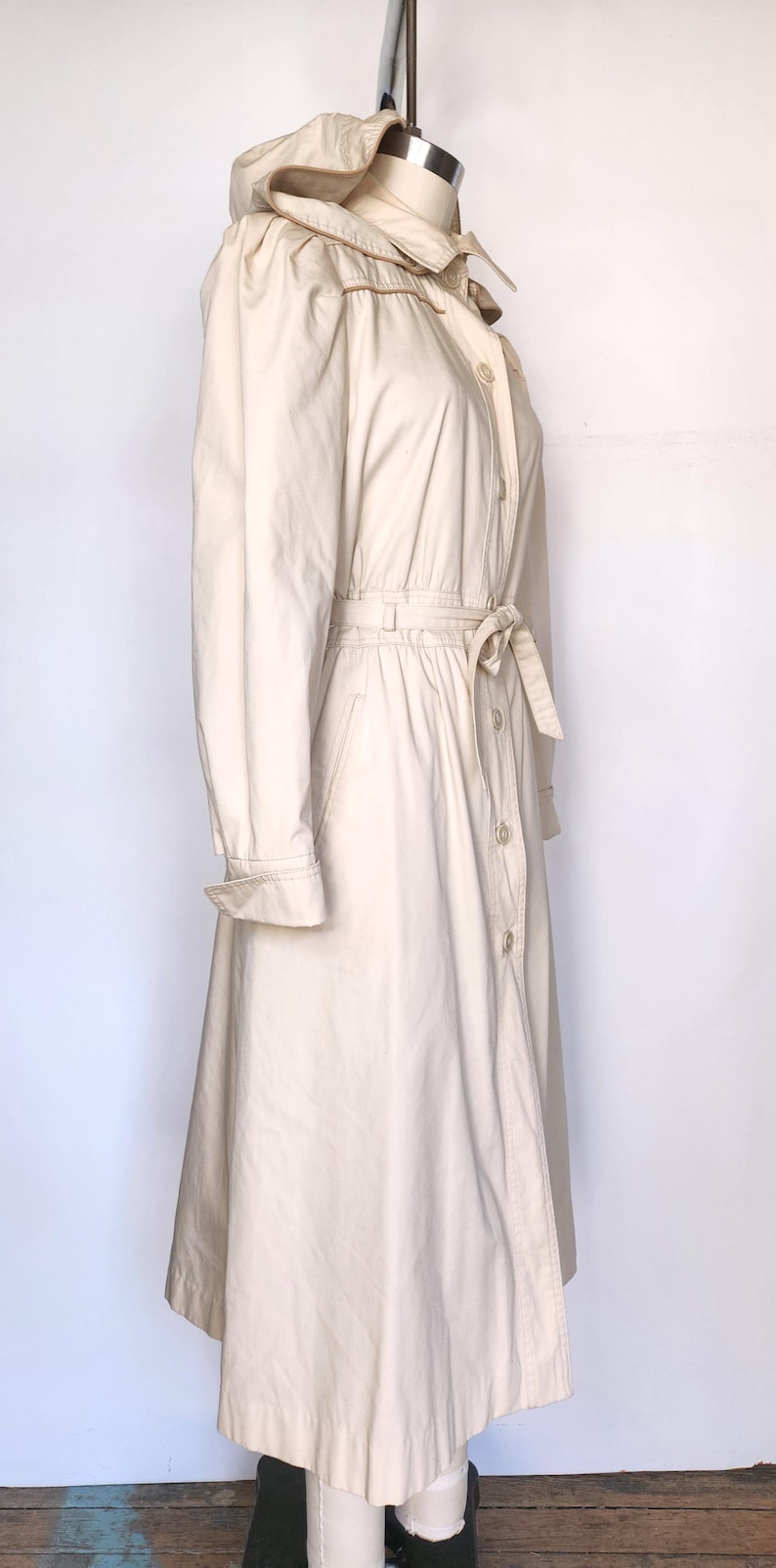 May include: A long, off-white hooded trench coat with a belt and button closure. The coat has a slightly flared skirt and a relaxed fit.