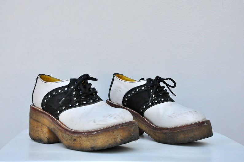 May include: A pair of white and black saddle shoes with thick platform soles. The shoes have a black leather trim and white laces.