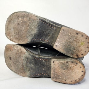 May include: Close-up of the soles of a pair of black leather boots. The soles are worn and have a pattern of small, round studs.