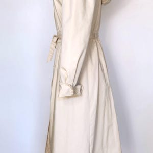 May include: A cream-colored hooded trench coat with a belt. The coat has a long, flowing silhouette and a relaxed fit.