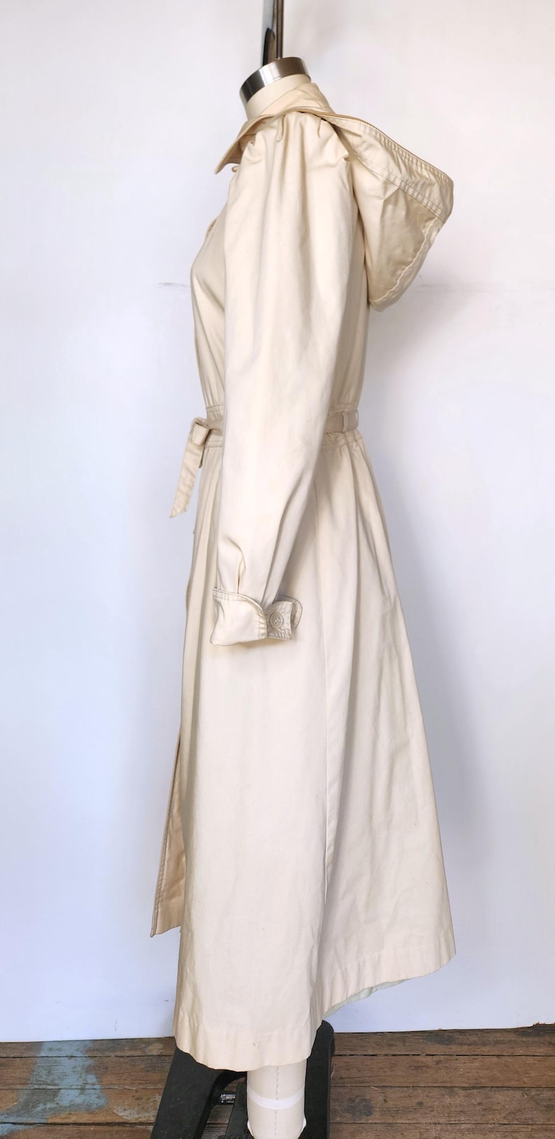 May include: A cream-colored hooded trench coat with a belt. The coat has a long, flowing silhouette and a relaxed fit.
