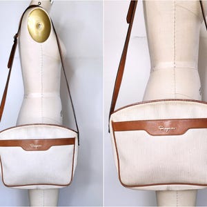 May include: A cream-colored shoulder bag with a textured fabric body and brown leather trim. The bag features a brown leather horizontal strap across the front with the gold-toned text Ferragamo embossed on it and a long adjustable shoulder strap.