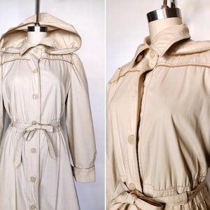 May include: A beige hooded trench coat with a button-up front, a tie belt, and long sleeves. The coat has a collar and a slightly flared skirt.