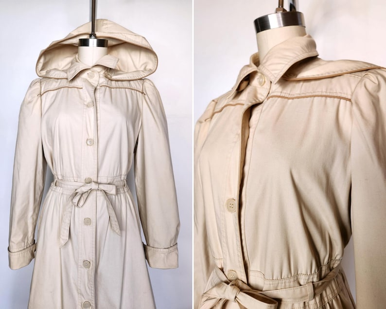 May include: A beige hooded trench coat with a button-up front, a tie belt, and long sleeves. The coat has a collar and a slightly flared skirt.