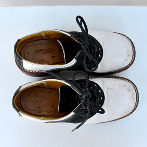 May include: A pair of white leather saddle shoes with black trim and laces. The shoes have a brown rubber sole.