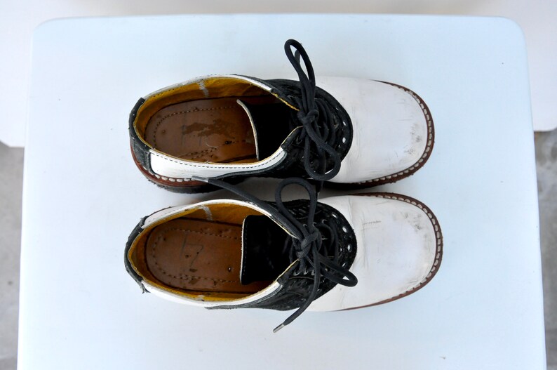 May include: A pair of white leather saddle shoes with black trim and laces. The shoes have a brown rubber sole.